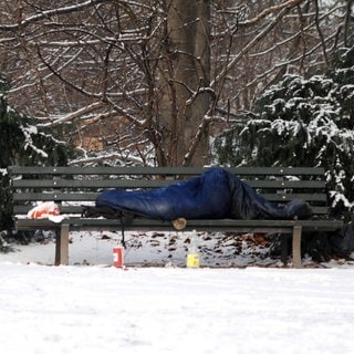 Ein Obdachloser liegt im Schlafsack auf einer Parkbank im Schnee.