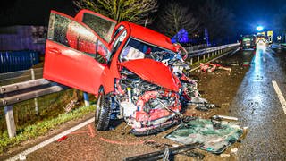 Das völlig zerstörte Wrack eines Autos liegt an einer Straße, auf der im Hintergrund die Rettungskräfte stehen.