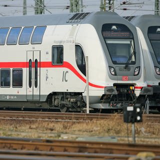 Zwei Doppelstockzüge der zweiten Generation des InterCity stehen auf den Gleisanlagen vor einemHauptbahnhof. 