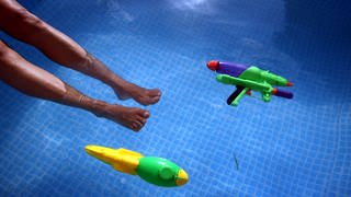 Füße baumeln im Wasser eines Swimming-Pools, daneben schwimmen zwei Wasser-Pistolen.