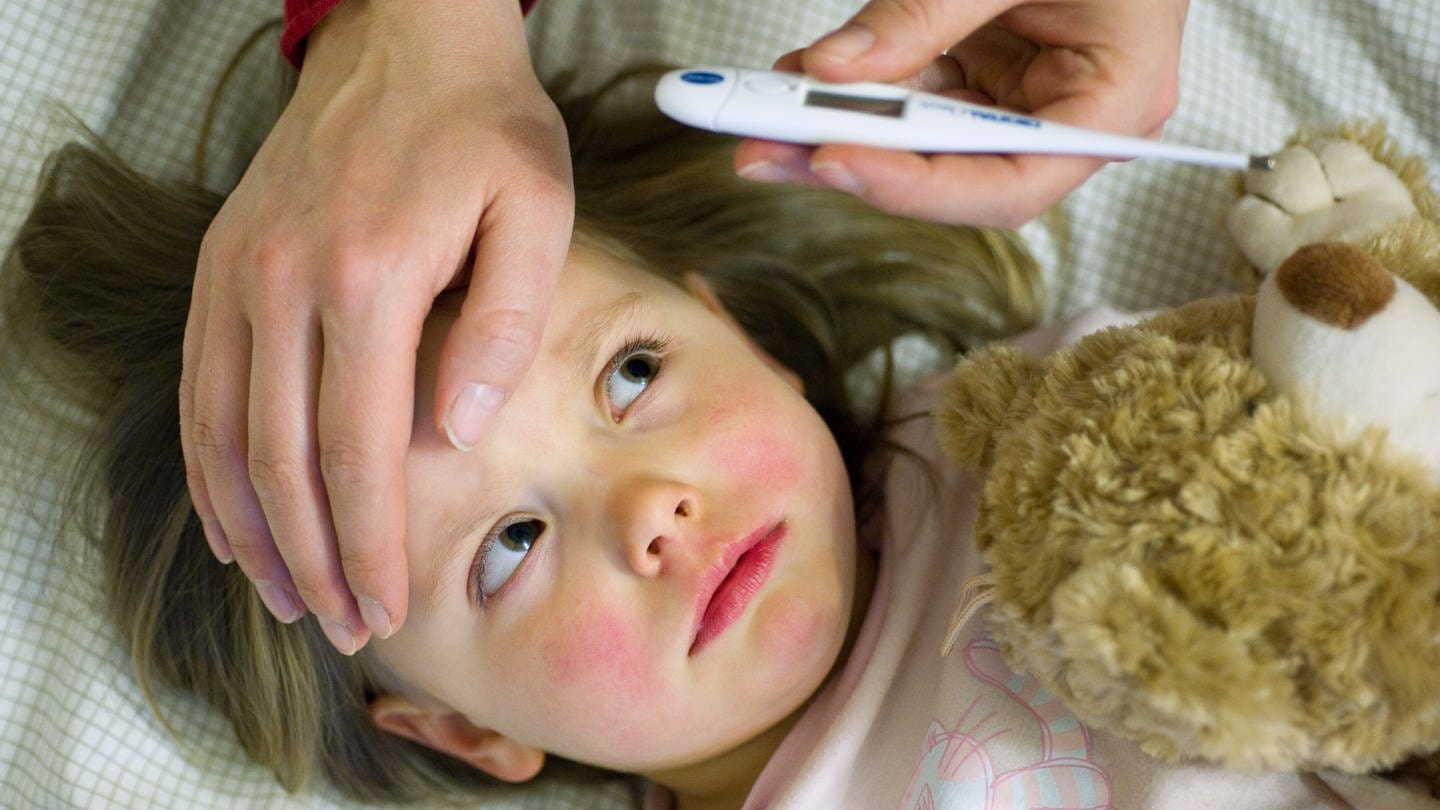 Ein krankes Kind liegt im Bett, die Mutter fühlt mit der Hand die Stirn und hält ein Thermometer in der Hand.