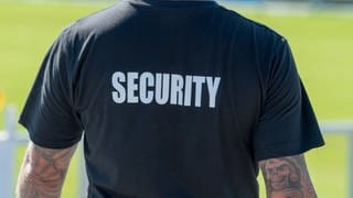 Ein Mann in einem T-Shirt mit der Aufschrift "Security" steht mit dem Rücken zur Kamera