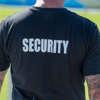 Ein Mann in einem T-Shirt mit der Aufschrift "Security" steht mit dem Rücken zur Kamera