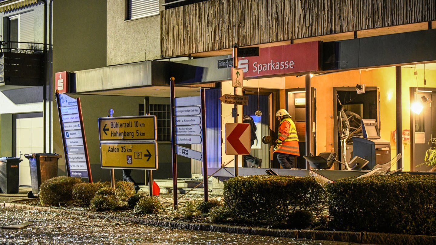 Die Bankfiliale von außen: Schilder wurden durch die Explosion verbogen, Glas liegt auf der Straße, Einsatzkräfte sind im Inneren der Filiale