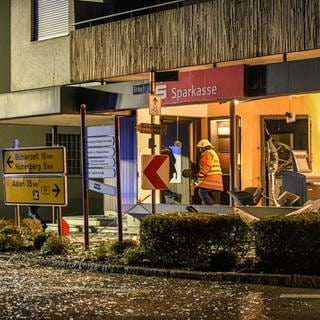 Die Bankfiliale von außen: Schilder wurden durch die Explosion verbogen, Glas liegt auf der Straße, Einsatzkräfte sind im Inneren der Filiale