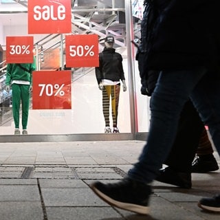 Personen gehen an einem Schaufenster mit der Aufschrift "Sale" und Preisreduktionen vorbei.