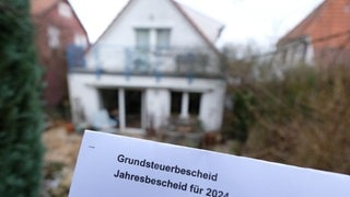 Ein Grundsteuerbescheid der Stadt für das Jahr 2024 wird vor einem Grundstück und einem Haus vor die Kamera gehalten.