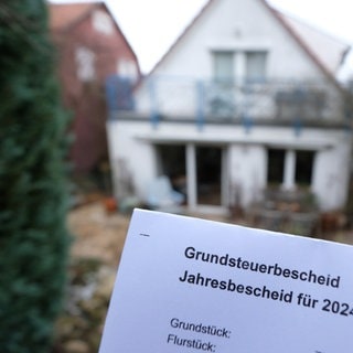 Ein Grundsteuerbescheid der Stadt für das Jahr 2024 wird vor einem Grundstück und einem Haus vor die Kamera gehalten.