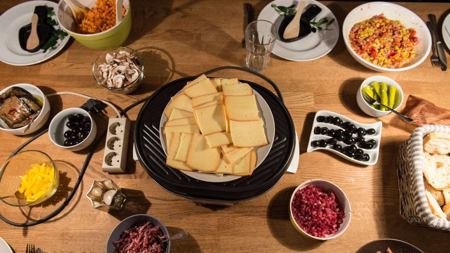 Raclettekäse und Schalen mit Zutaten stehen auf einem Tisch.