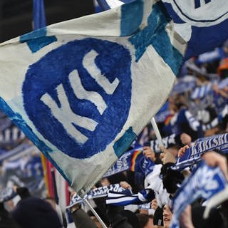 Eine KSC-Fahne im Vordergrund, im Hintergund unscharf KSC-Fans mit Schals auf der Tribüne.