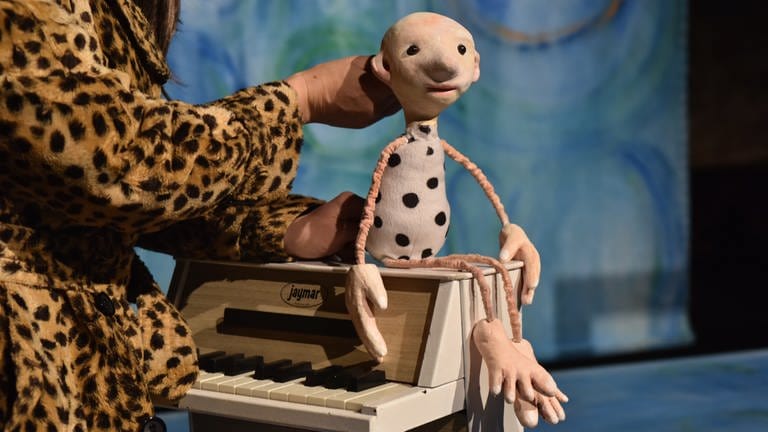 Eine stilisierte Puppe mit dünnen Ärmchen und Beinchen und einenm birnenförmigen Körper mit schwarzen Punkten, sitzt af einem Miniatur-Klavier.