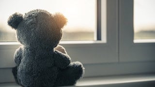 Symbolbild: Ein Teddybär sitzt am Fenster. Kinder die vor, während oder kurz nach der Geburt sterben, nennt man auch Sternenkinder. Eine Trauerbegleitung kann betroffene Eltern bei dem Verlust unterstützen. 