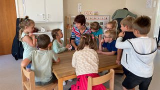 Viele Kinder sitzen rund um einen Tisch zusammen mit einer Erzieherin