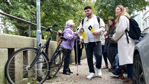 Christos Intos hängt eine Gelbe Karte an ein Fahrrad auf dem Gehweg. Im Hintergrund erklärt Brigitte Schick den Schülern die Verletzungsgefahr.