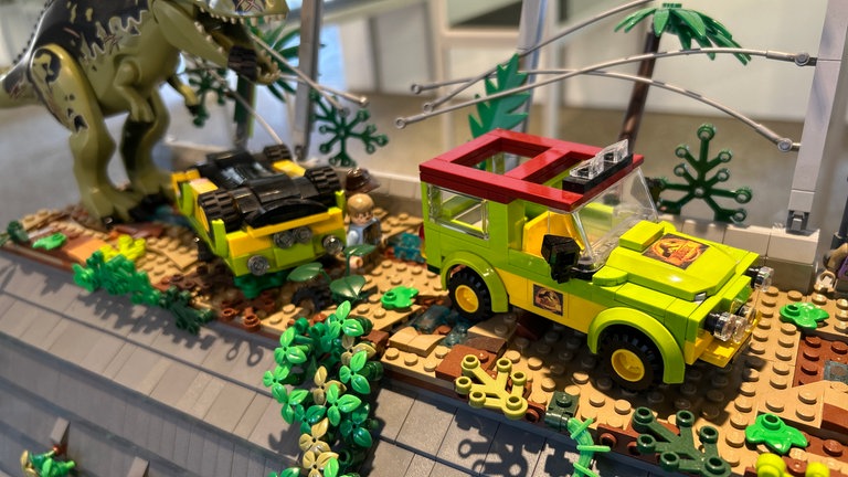 In der Lego-Ausstellung in Karlsruhe durchbricht ein Dino einen Zaun und greift zwei Autos an.