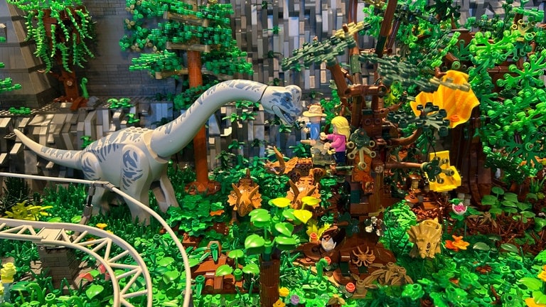 Ein Langhals-Dinosaurier nähert sich mit seinem Kopf drei Lego-Figuren, die sich auf einen Baum in einer Dschungel-Landschaft gerettet haben.