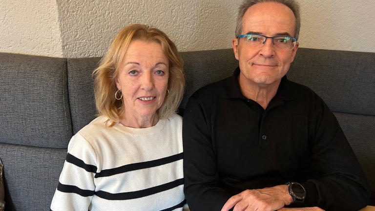 Eine Frau mit Ohrringen und ein Mann mit Brille sitzen auf einer grauen Sitzbank.