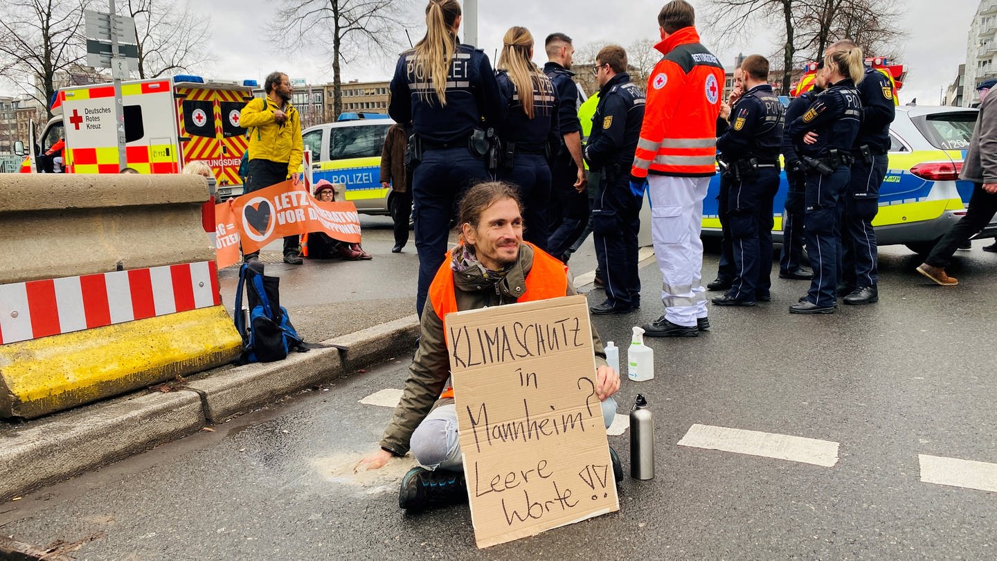 Klimaaktivist sitzt mit Plakat auf Straße. Eine Hand ist auf Straße festgeklebt.