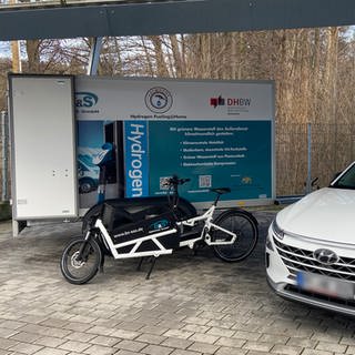 Ein Wasserstoff betriebenes Auto steht neben dem Prototyp eienr Wasserstofftankstelle.