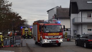 Zahlreiche Einsatzkräfte sind vor Ort. Ein Einsatzfahrzeug der Feuerwehr steht auf der Straße, im Hintergrund wird durch ein Fenster unter dem Dach gelöscht.