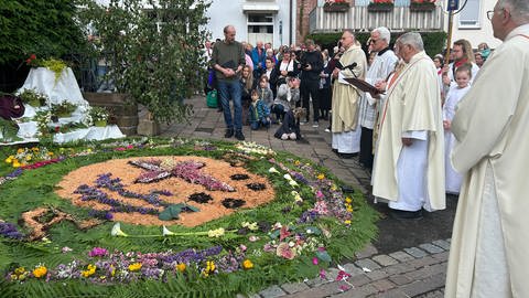 Geistliche und Pilger stehen an einem Blumenteppich bei der Fronleichnamsprozession in Walldürn