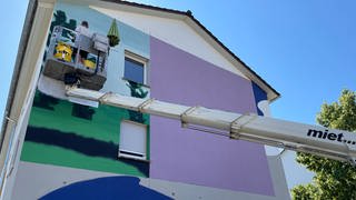 Der Künstler Perkup steht auf einer Hebebühne. Er hat die Hauswand in Blau- und Fliedertönen grundiert und angefangen, eine Landschaft darauf zu malen 