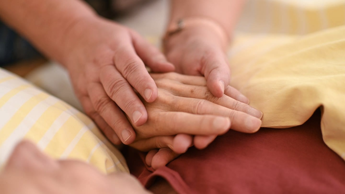 Eine Hospizmitarbeiterin hält die Hand eines todkranken Menschen, der in einem Hospiz im Bett liegt