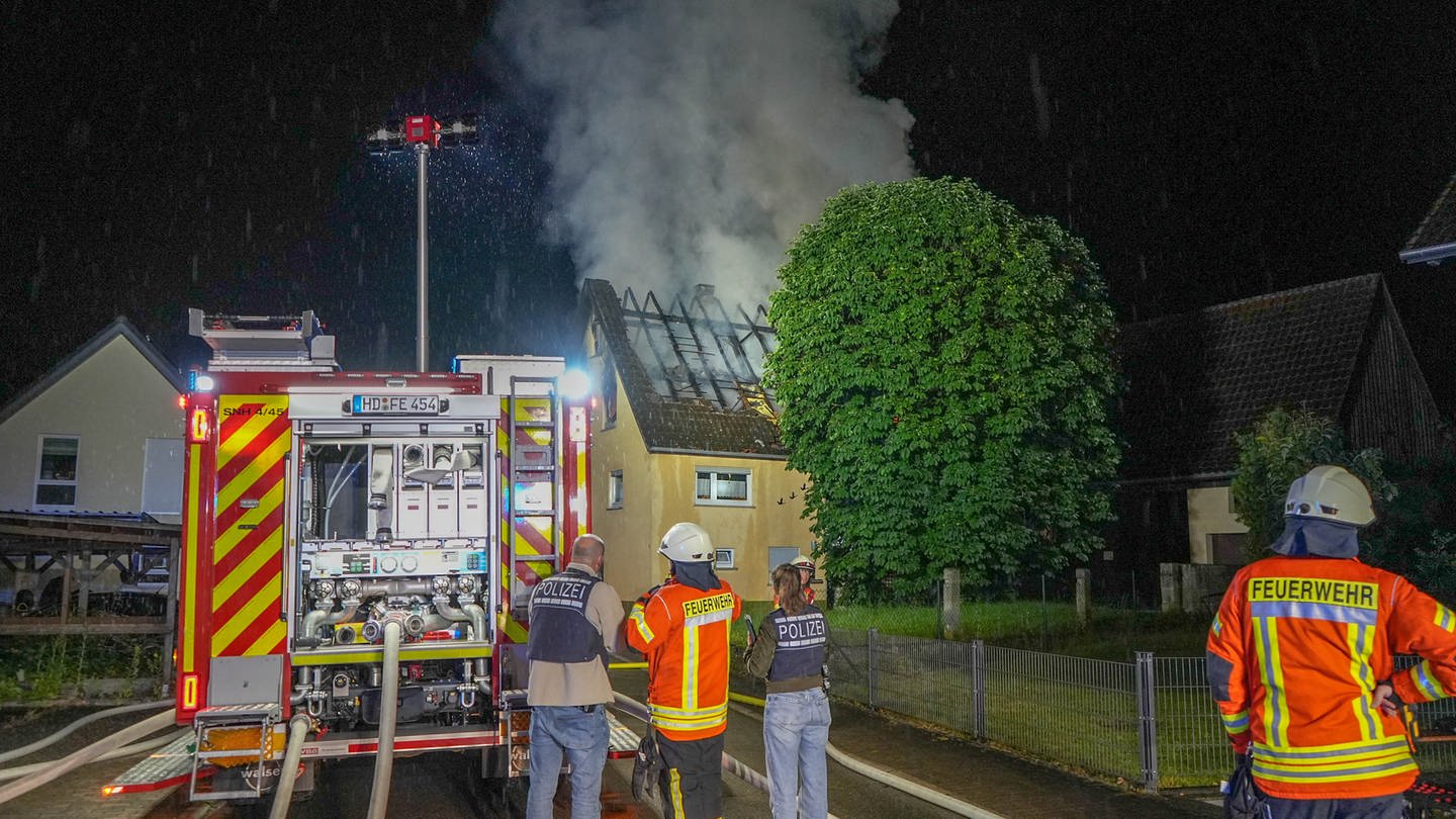 Rettungskräfte stehen neben einem Feuerwehr-Fahrzeug in Sinsheim-Ehrstädt (Rhein-Neckar-Kreis). Dahinter steigt Rauch aus einem Wohnhaus auf.