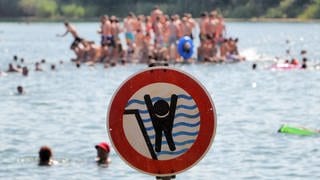 Badesee mit Badegästen. Ein Schild warnt vor einem stark abfallenden Ufer.