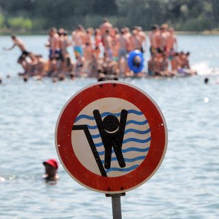 Badesee mit Badegästen. Ein Schild warnt vor einem stark abfallenden Ufer.