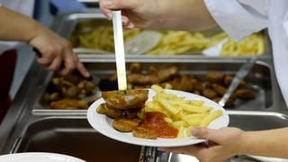 Symbolbild: Essen wird in Kantine ausgegeben - Mensch mit Schöpfkelle gießt Soße über Pommes und Frikadellen