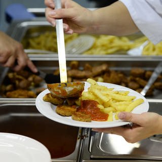 Symbolbild: Essen wird in Kantine ausgegeben - Mensch mit Schöpfkelle gießt Soße über Pommes und Frikadellen