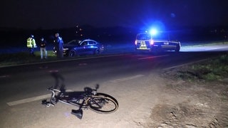 Auf einer Kreisstraße bei Heddesheim (Rhein-Neckar-Kreis) liegt nach einem Unfall ein Fahrrad, im Hintergrund neben der Straße ein Auto und Polizisten, rechts ein Polizeifahrzeug.