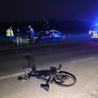 Auf einer Kreisstraße bei Heddesheim (Rhein-Neckar-Kreis) liegt nach einem Unfall ein Fahrrad, im Hintergrund neben der Straße ein Auto und Polizisten, rechts ein Polizeifahrzeug.