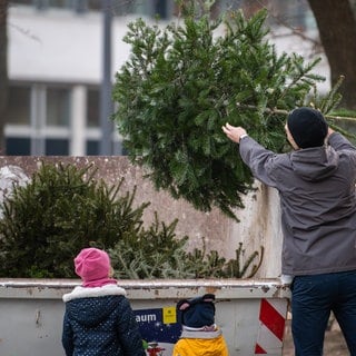 Ein Mann wirft vor seinen Kindern einen alten Weihnachtsbaum in einen Container.