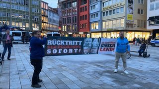Demonstranten der AfD stehen auf dem Stuttgarter Marktplatz und halten ein Banner.