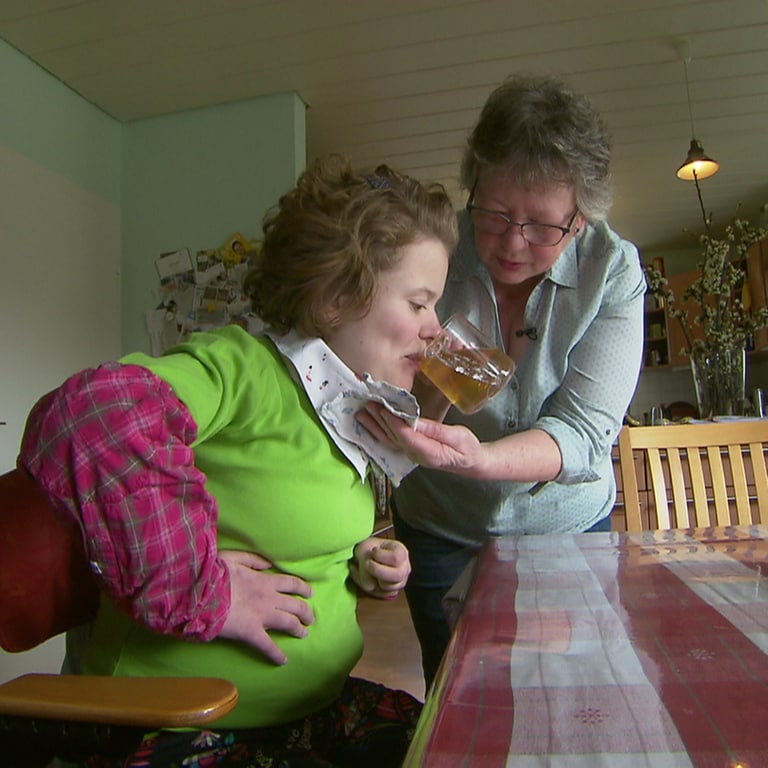 Ursula Hofmann kümmert sich seit 21 Jahren im ihre schwerbehinderte Tochter Anne. Die beiden sitzen am Esstisch und die Mutter gibt der Tochter aus einem Glas etwas zu trinken.