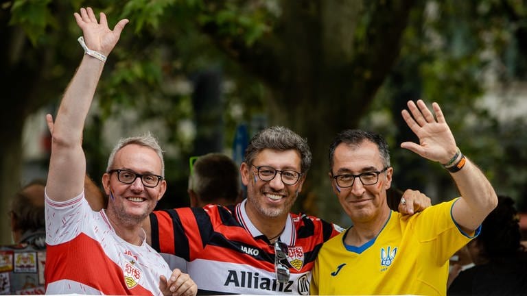 V.l.n.r.: Alexander Wehrle, Vorstandsvorsitzender VfB Stuttgart, VfB-Präsident Claus Vogt und Cem Özdemir (Grüne) winken, im Gesicht tragen sie regenbogenfarbene Schminke.