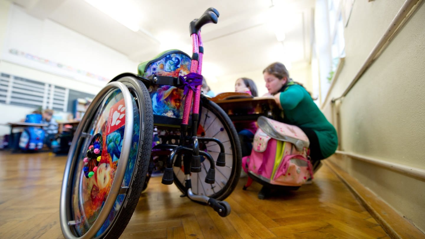 Eine junge Frau, die ein Freiwilliges Soziales Jahr (FSJ) macht, betreut in einem Klassenzimmer einer Gemeinschaftsschule in Stuttgart eine Grundschülerin, die auf einen Rollstuhl angewiesen ist (Archivbild).