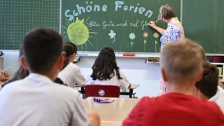 Schulkinder einer 4. Klasse einer Stuttgarter Grundschule sitzen im Klassenzimmer am letzten Schultag vor den Sommerferien in Baden-Württemberg während eine Lehrerin an eine Tafel "Schöne Ferien Alles Gute und viel Glück" schreibt.