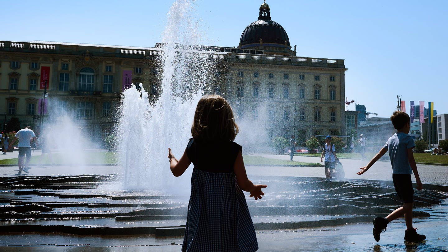 Bei wolkenlosem Himmel und sommerlichen Temperaturen am Lustgarten spielen Kinder am Brunnen.