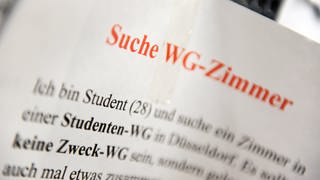  Ein Zettel mit der Aufschrift "Suche WG-Zimmer" hängt an einem Schwarzen Brett.