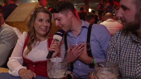 Reporterin Kristin Haub sitzt in einem Festzelt auf dem Wasen und ist von Bier trinkenden Gästen umgegeben.