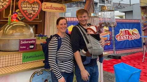 Wasen 2024: Eva und Max sind große Volksfest-Fans. Ihrer kleinen Tochter haben sie extra eine Baby-Lederhose angezogen und wollen ihr ein Lebkuchenherz hier am Stand kaufen.