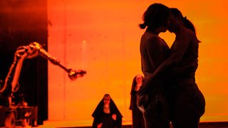 Zwei Frauen umarmen sich bei der Aufführung der Opernperformance "Sancta", während im Hintergrund Nonnen zu sehen sind.