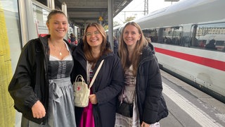 Auf einem Bahnsteig am Stuttgarter Hauptbahnhof stehen Gina, Paula und Sophia und schauen lächelnd in die Kamera.