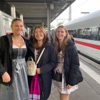 Auf einem Bahnsteig am Stuttgarter Hauptbahnhof stehen Gina, Paula und Sophia und schauen lächelnd in die Kamera.