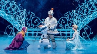 Anna und Elsa knien im Musical "Die Eiskönigin" in Stuttgart vor dem Schneemann Olaf.