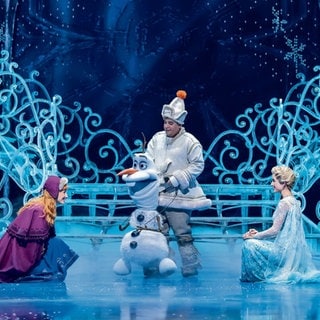 Anna und Elsa knien im Musical "Die Eiskönigin" in Stuttgart vor dem Schneemann Olaf.