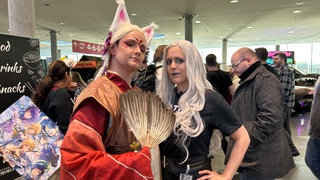 Zwei junge Frauen in aufwendigen Kostümen schauen auf der Comic Con Stuttgart in die Kamera. 